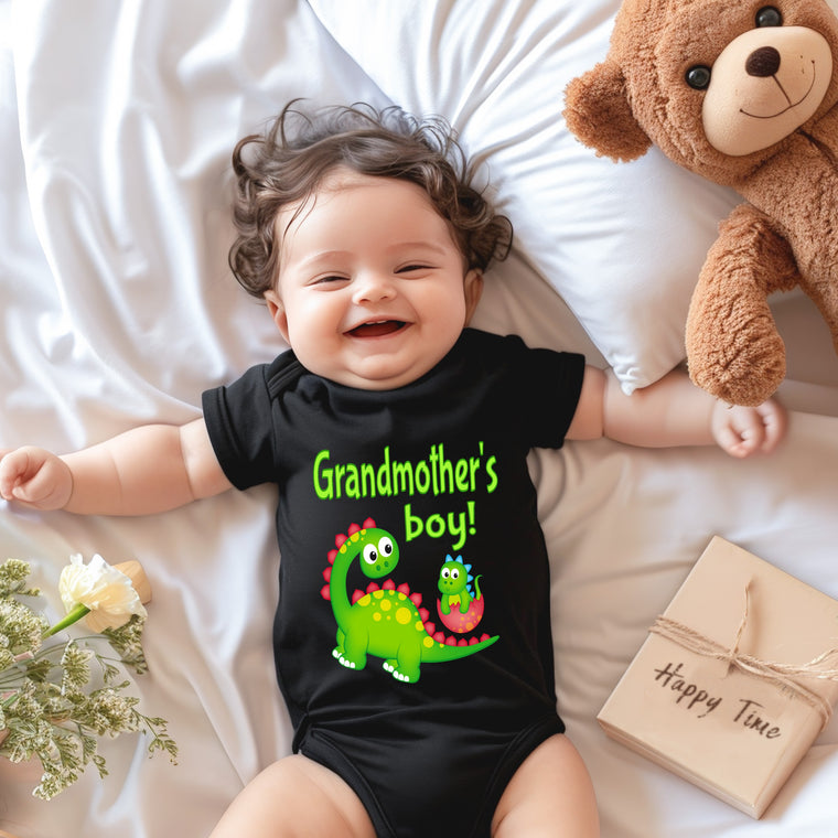 A smiling baby wearing a black t-shirt with a dinosaur graphic lies on a bed next to a stuffed teddy bear.