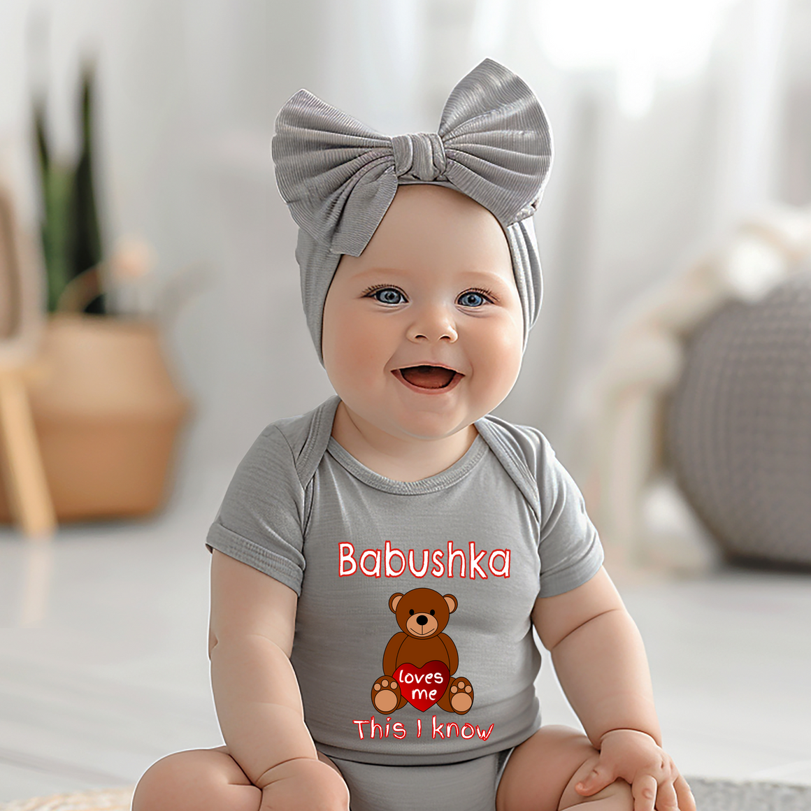 A smiling baby girl wearing a gray headband and a gray onesie with a teddy bear graphic, sitting on a white floor.