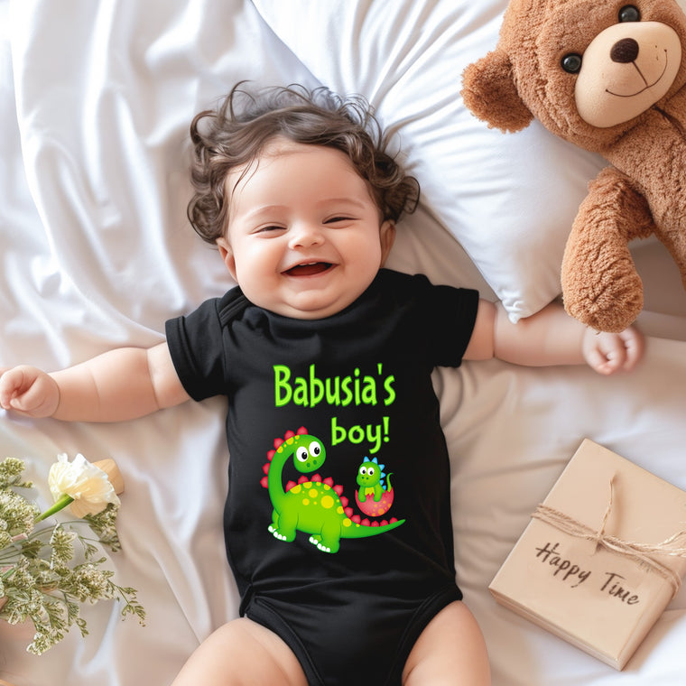 A smiling baby wearing a black t-shirt with a dinosaur graphic lies on a bed next to a stuffed teddy bear.