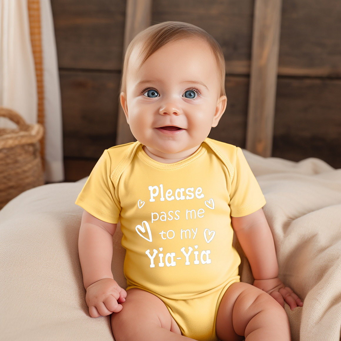 A smiling baby wearing a yellow onesie with the text "Please pass me to my Yia-Yia" sits on a bed.