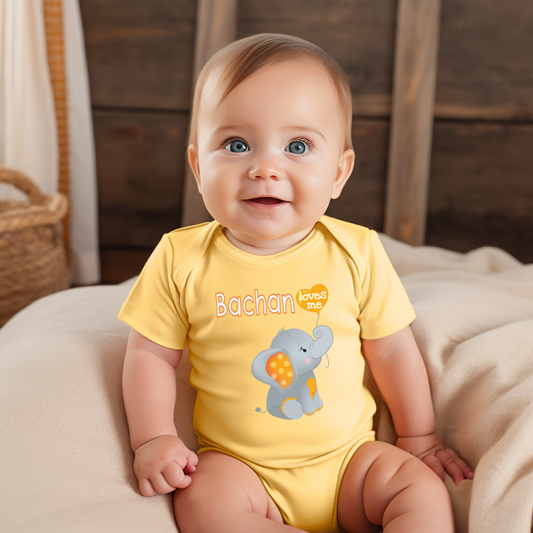 A smiling baby boy wearing a yellow onesie with an elephant design sits on a bed.