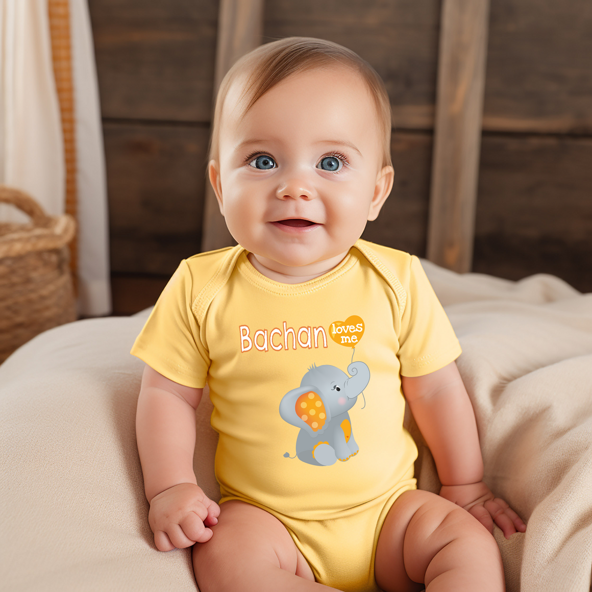 A smiling baby boy wearing a yellow onesie with an elephant design sits on a bed.