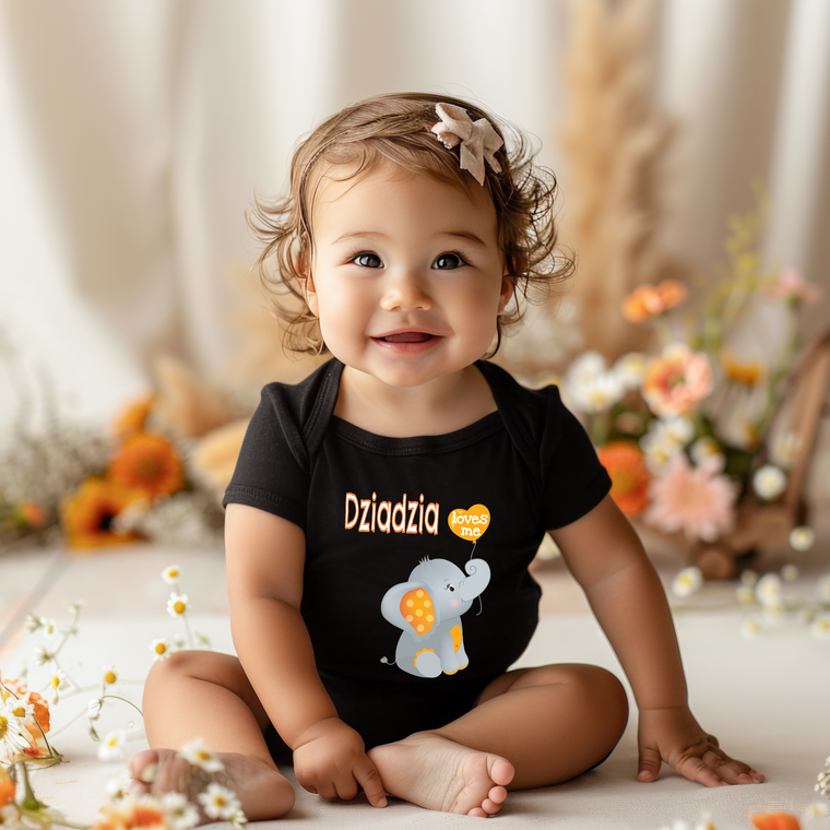 A smiling baby girl wearing a black shirt with an elephant design sits on the floor surrounded by flowers.
