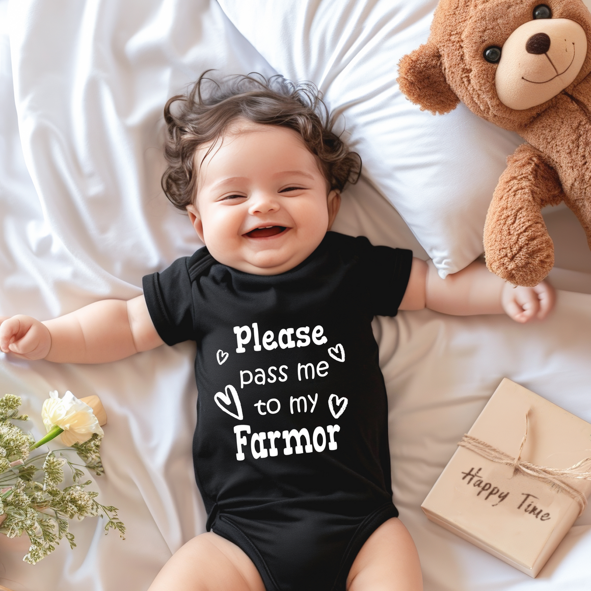 A smiling baby wearing a black onesie with the text "Please pass me to my Farmer" lies on a bed next to a teddy bear and a bouquet of flowers.