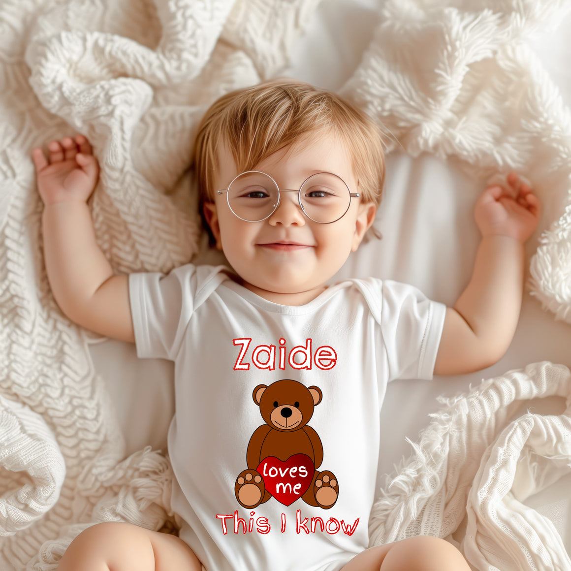A smiling baby wearing a white onesie with a teddy bear graphic and the text "Zaide loves me This I know" lying on a white blanket.