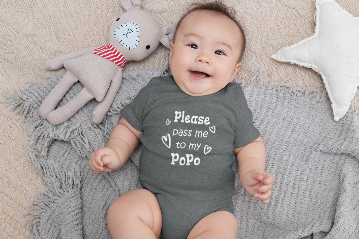 A smiling baby wearing a gray onesie with the text "Please pass me to my Popo" lies on a gray blanket next to a stuffed animal.