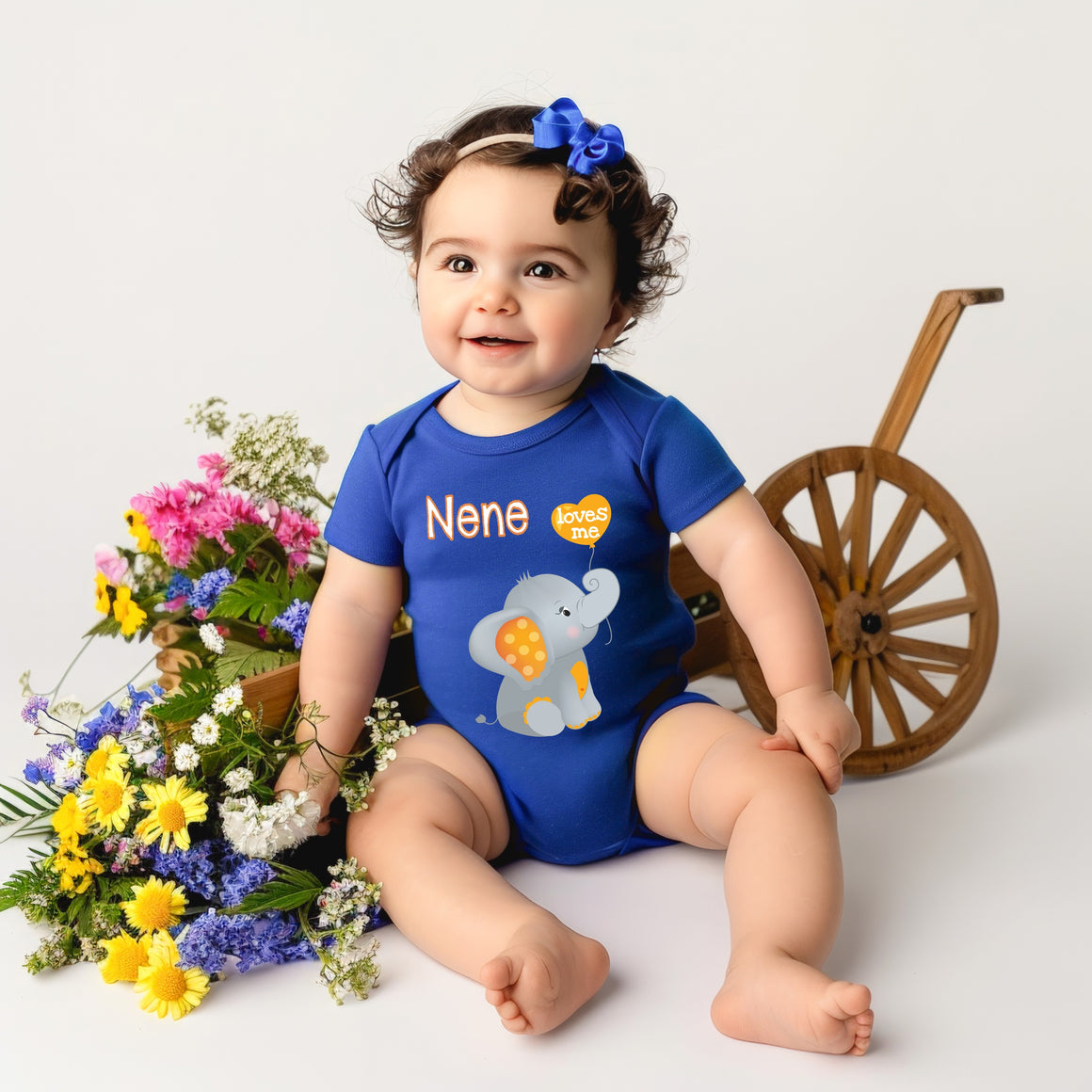 A smiling baby girl wearing a blue onesie with an elephant design sits next to a wooden wagon filled with colorful flowers.