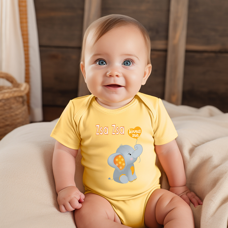 A smiling baby wearing a yellow onesie with an elephant design sits on a bed.