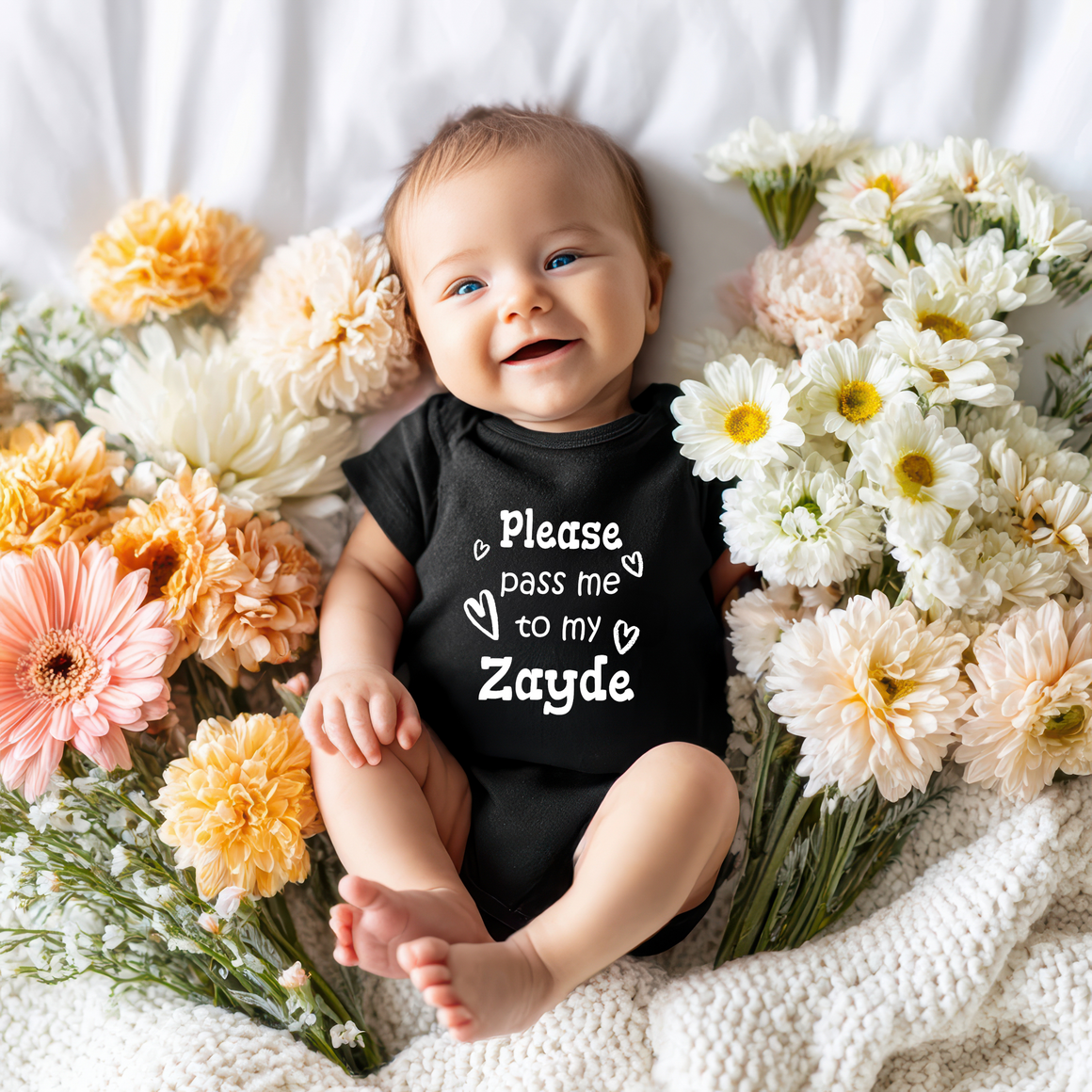 A smiling baby wearing a black outfit with the text "Please pass me to my Zayde" is surrounded by a bouquet of colorful flowers.