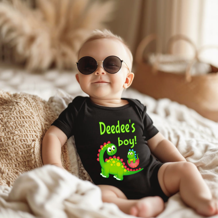 A baby wearing a black shirt with a dinosaur graphic and sunglasses is sitting on a bed.