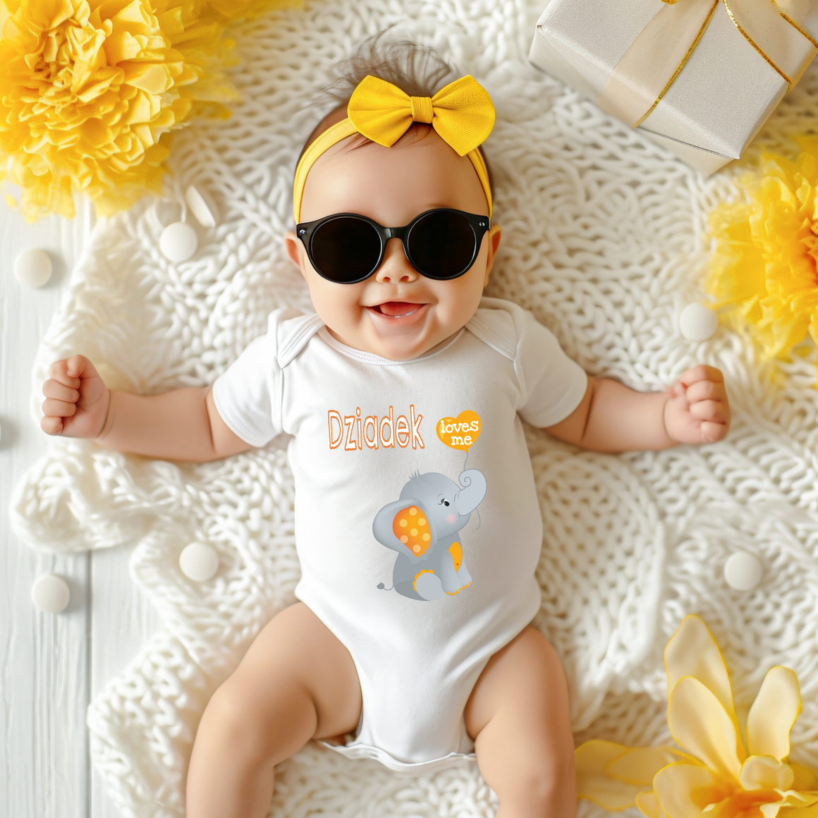 A baby wearing sunglasses and a yellow bow is lying on a white blanket surrounded by yellow flowers.