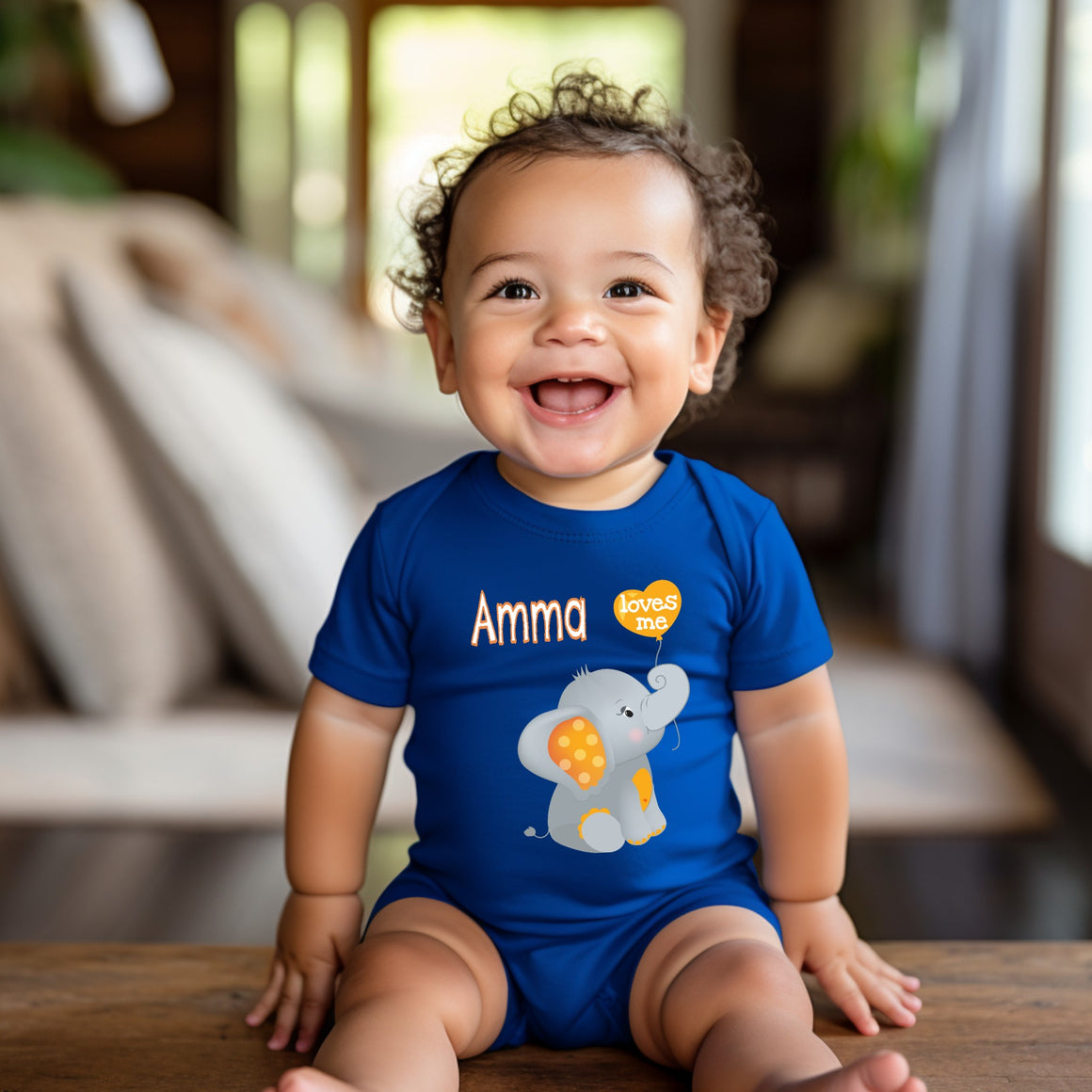 A smiling baby boy wearing a blue onesie with an elephant design sits on a wooden surface.