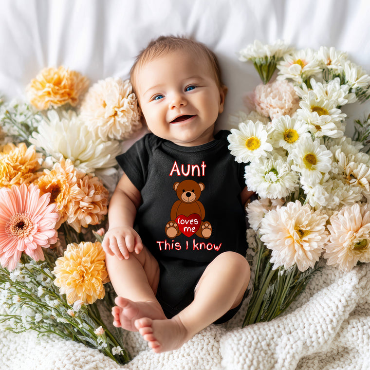 A smiling baby wearing a black shirt with a teddy bear graphic lies on a bed surrounded by colorful flowers.