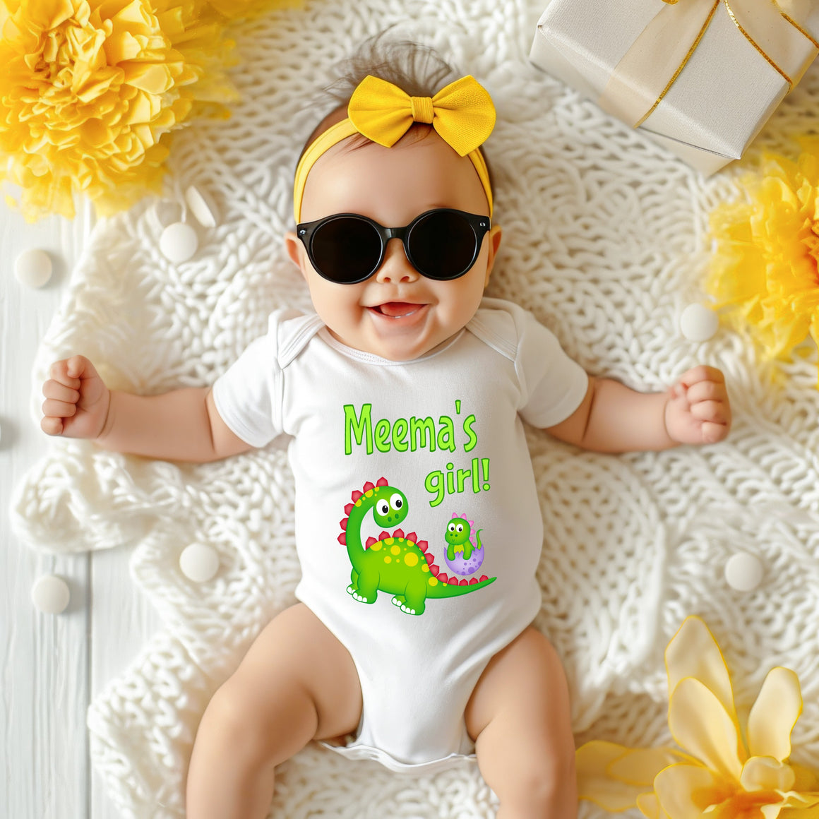 A baby wearing a white onesie with a dinosaur design and sunglasses lying on a white blanket surrounded by yellow flowers.