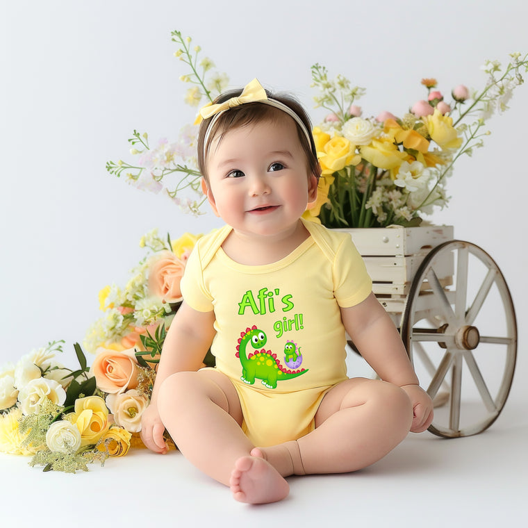 A smiling baby girl wearing a yellow onesie with a dinosaur graphic sits in front of a wooden wheel and a basket filled with yellow and white flowers.