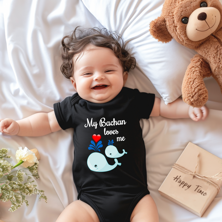 A smiling baby wearing a black onesie with a whale design lies on a bed next to a stuffed teddy bear.