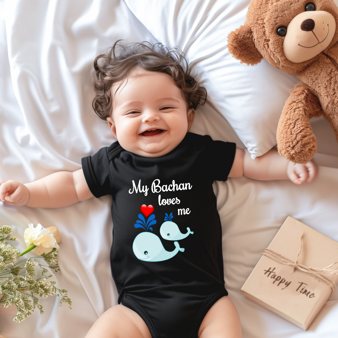 A smiling baby wearing a black onesie with a whale design lies on a bed next to a stuffed teddy bear.