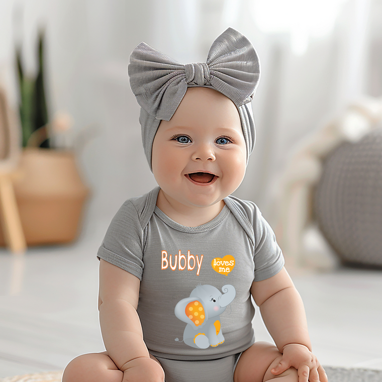 A smiling baby girl wearing a gray headband and a gray onesie with an elephant design, sitting on a white floor.