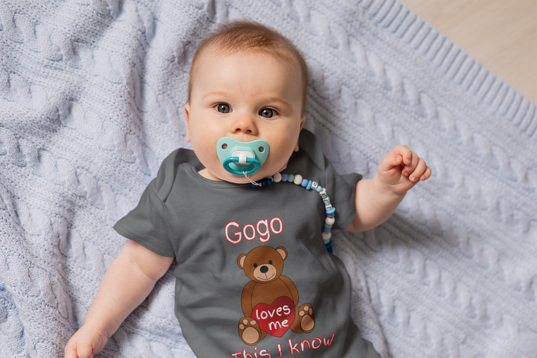 A baby wearing a gray t-shirt with a teddy bear graphic and the text "Gogo loves me" lying on a blue blanket.