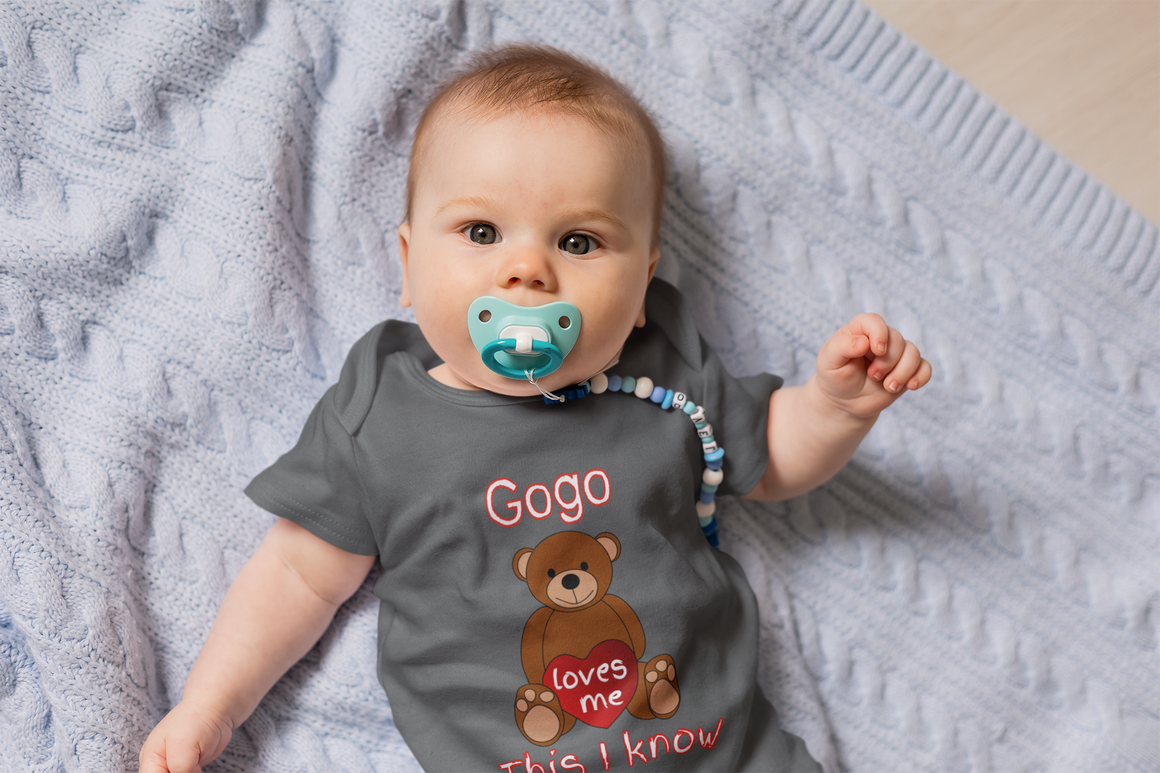 A baby wearing a gray t-shirt with a teddy bear graphic and the text "Gogo loves me" lying on a blue blanket.