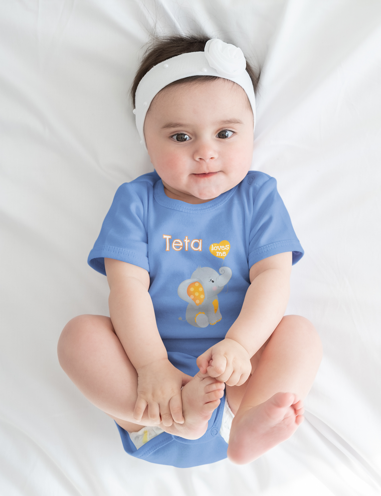 A baby girl wearing a blue onesie with an elephant design is lying on a white bed, smiling at the camera.