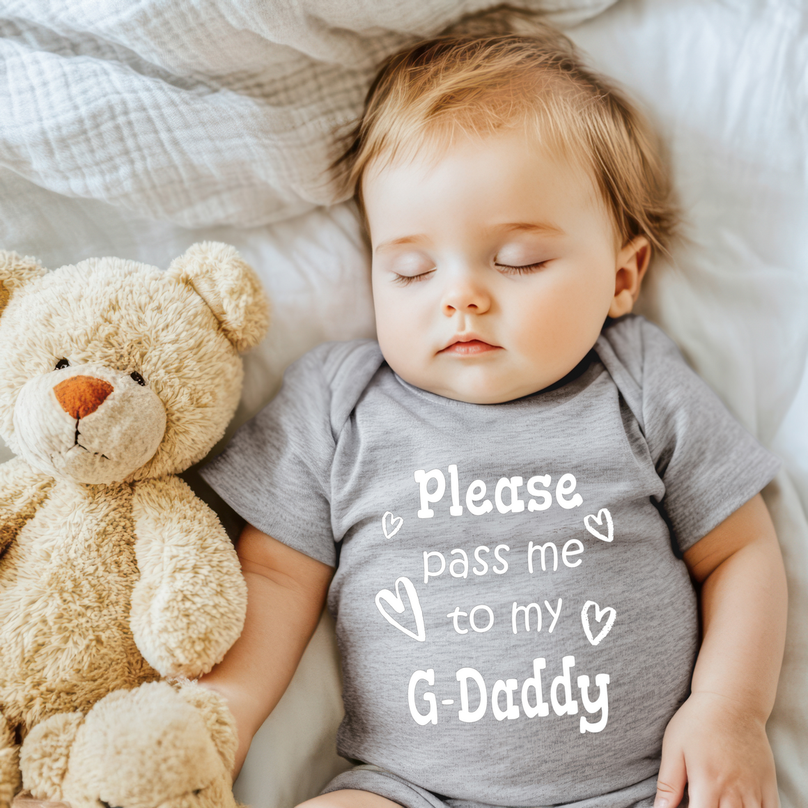 A sleeping baby wearing a gray onesie with the text "Please pass me to my G-daddy" is lying next to a teddy bear on a bed.