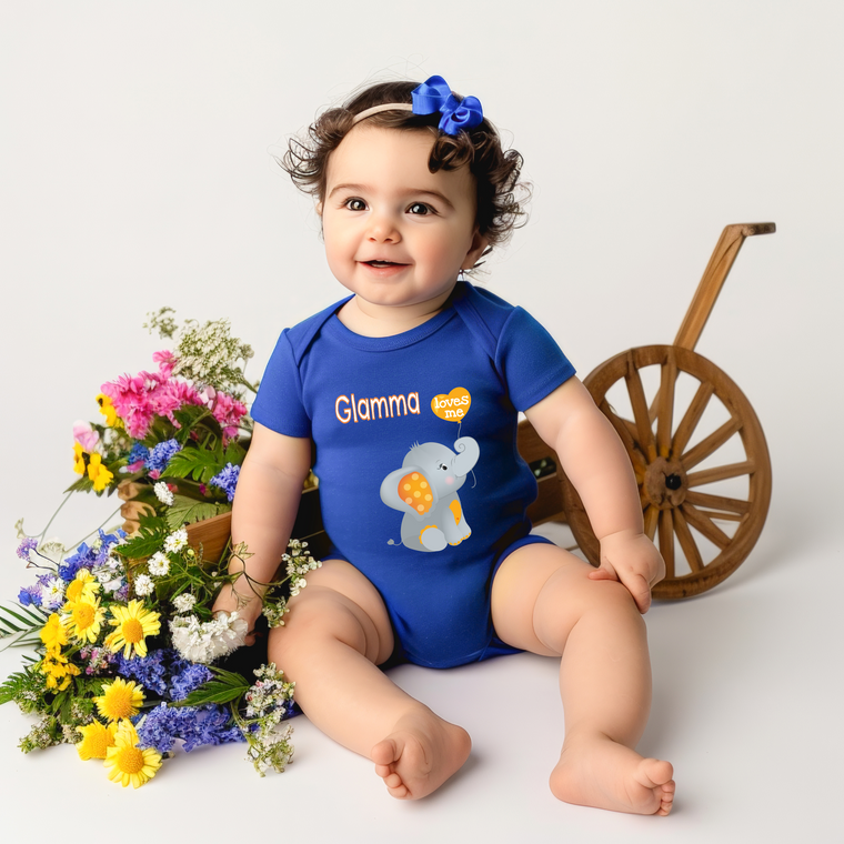 A smiling baby girl wearing a blue onesie with an elephant design sits next to a wooden wagon filled with colorful flowers.