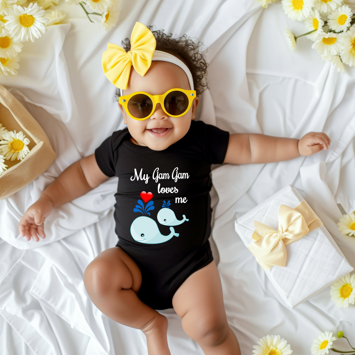 A smiling baby girl wearing a black outfit with a whale design and yellow sunglasses lies on a white sheet, surrounded by yellow flowers.