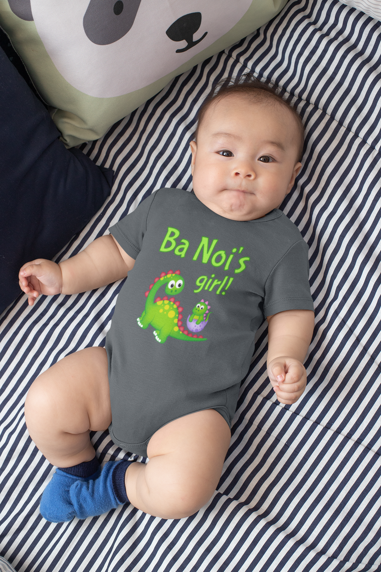 A baby wearing a grey onesie with a green dinosaur and the text "Ba Noi's girl!" lying on a striped bed.