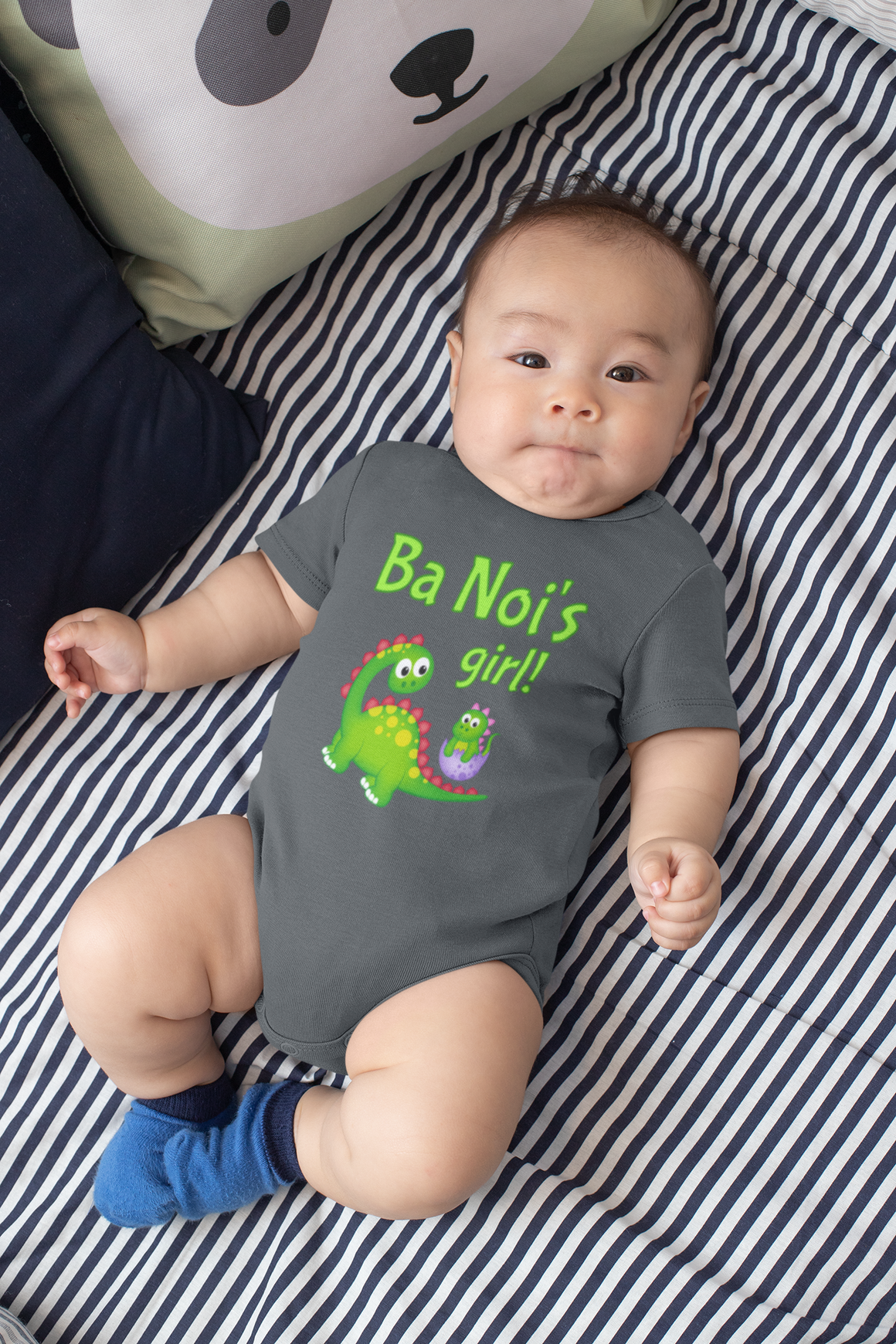 A baby wearing a grey onesie with a green dinosaur and the text "Ba Noi's girl!" lying on a striped bed.
