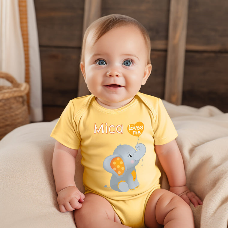A smiling baby boy wearing a yellow onesie with an elephant design sits on a bed.