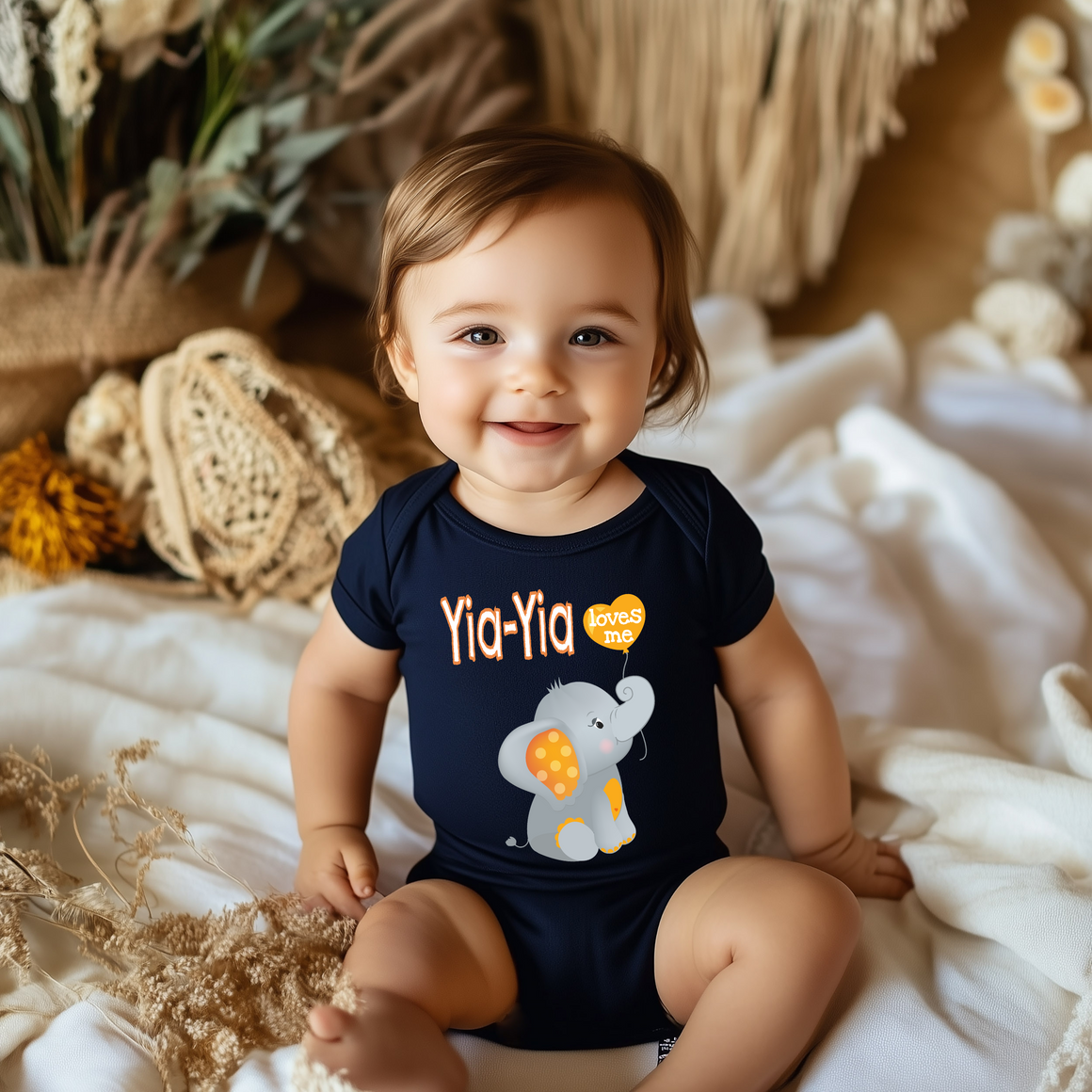 A smiling baby boy wearing a navy blue onesie with an elephant design sits on a white blanket.