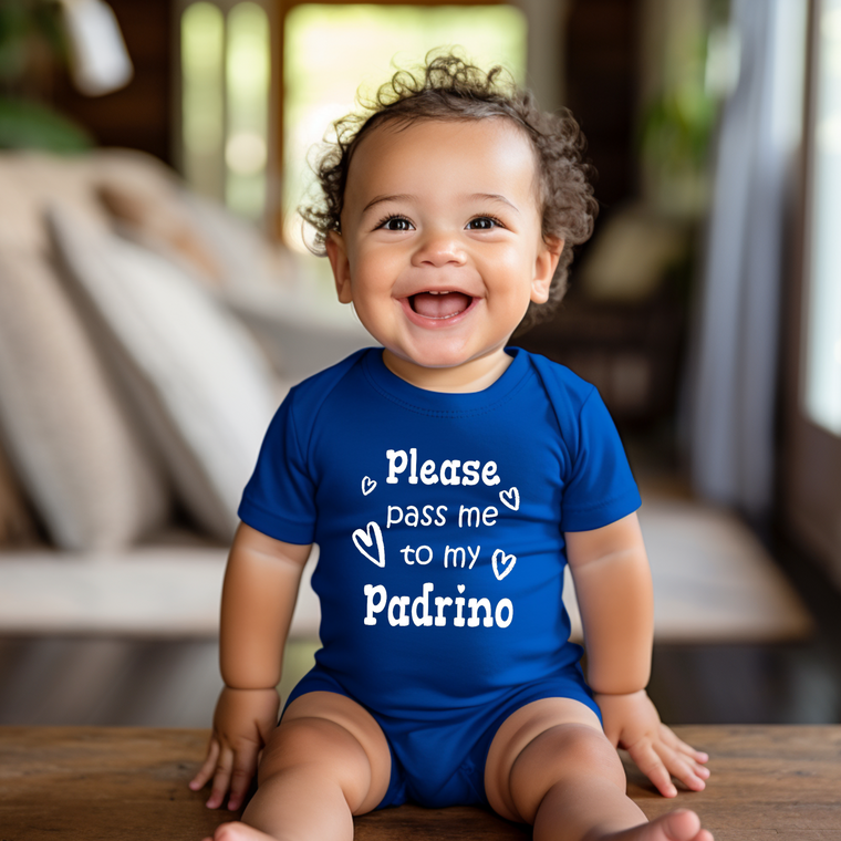 A smiling baby boy wearing a blue onesie with the text "Please pass me to my Padrino" is sitting on a wooden surface.