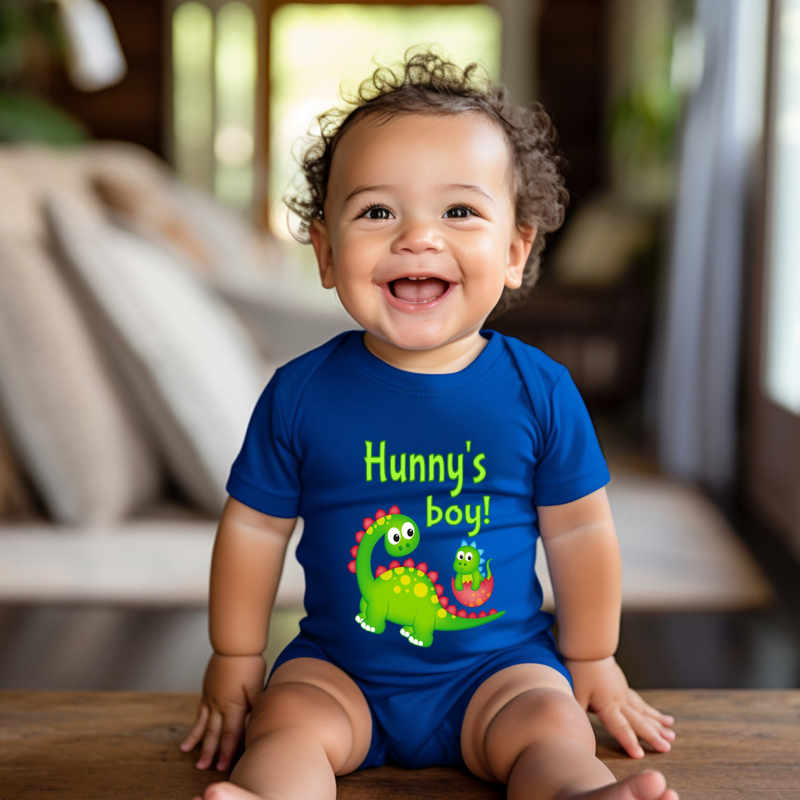 A smiling baby boy wearing a blue onesie with a dinosaur design and the text "Hunny's boy!"