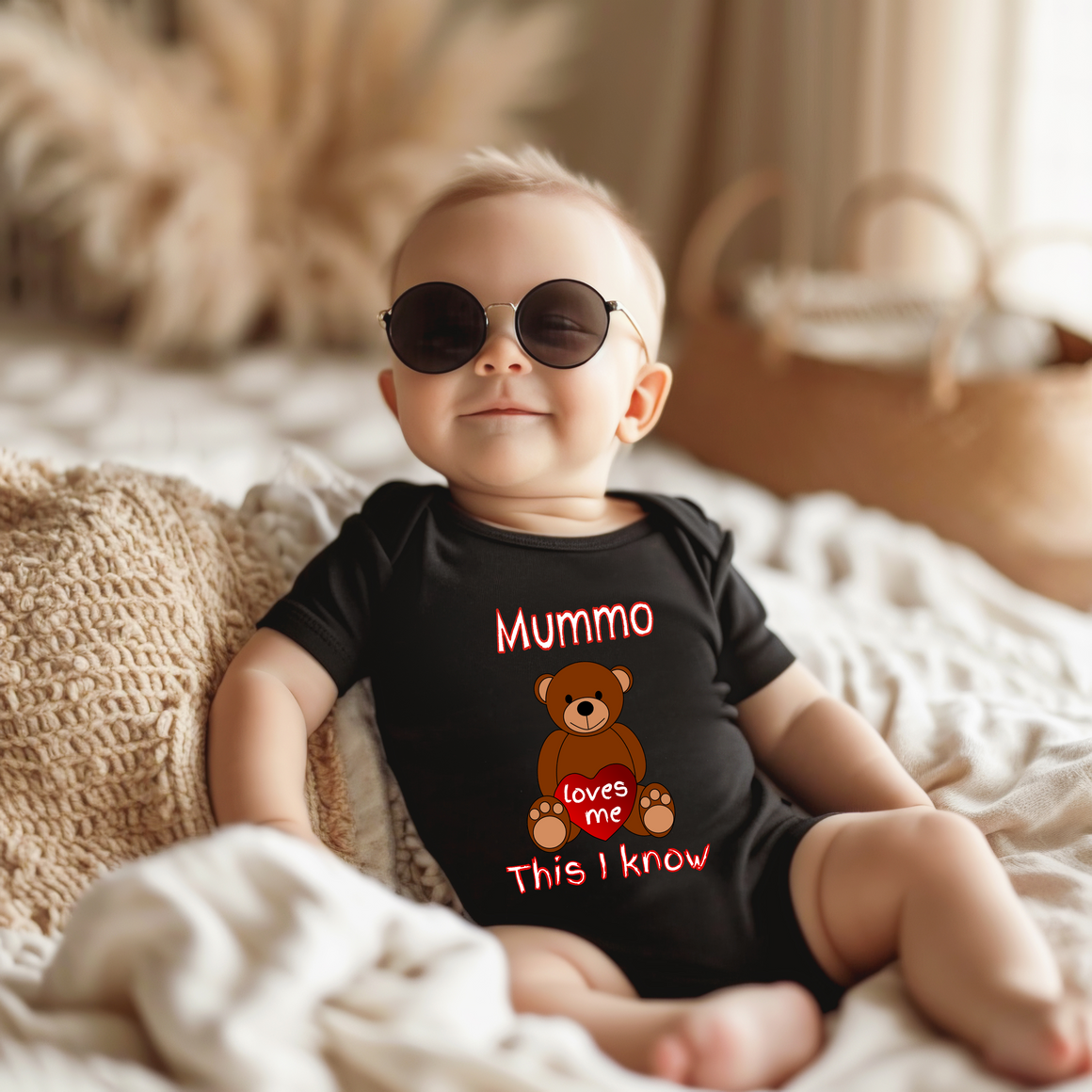 A baby wearing a black shirt with a teddy bear and the text "Mummo loves me" is sitting on a bed.