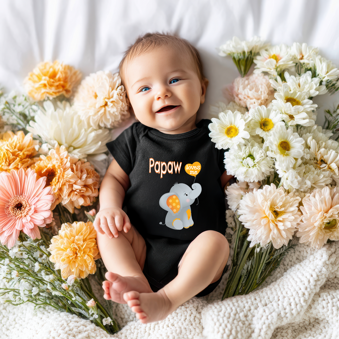 A smiling baby wearing a black onesie with an elephant design lies on a bed surrounded by colorful flowers.