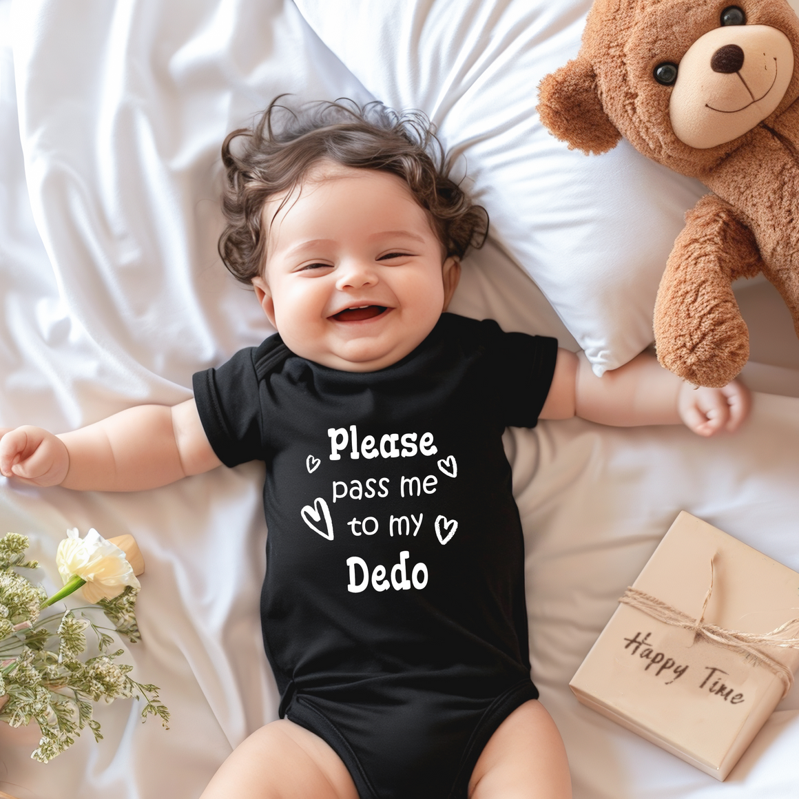 A smiling baby wearing a black onesie with the text "Please pass me to my Deco" lies on a bed next to a teddy bear and a gift box.