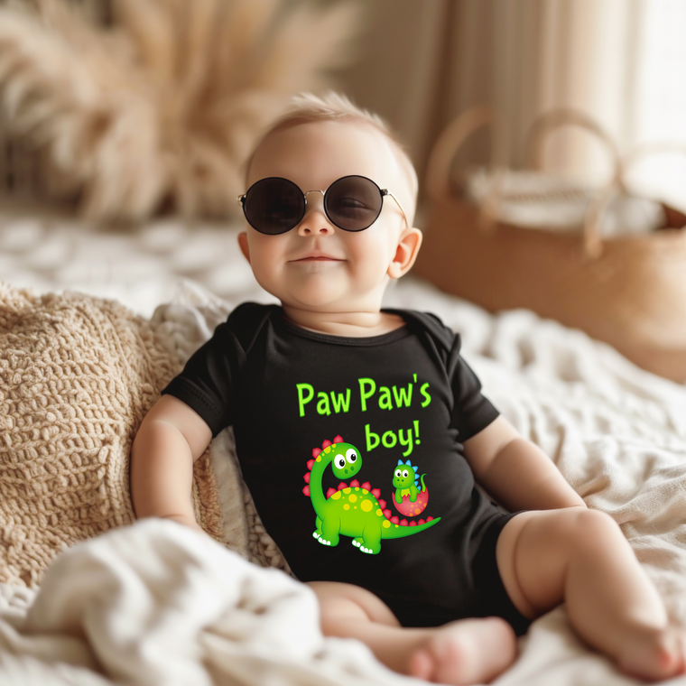 A baby wearing a black onesie with a green dinosaur and the text "Paw Paw's boy!" is sitting on a bed.