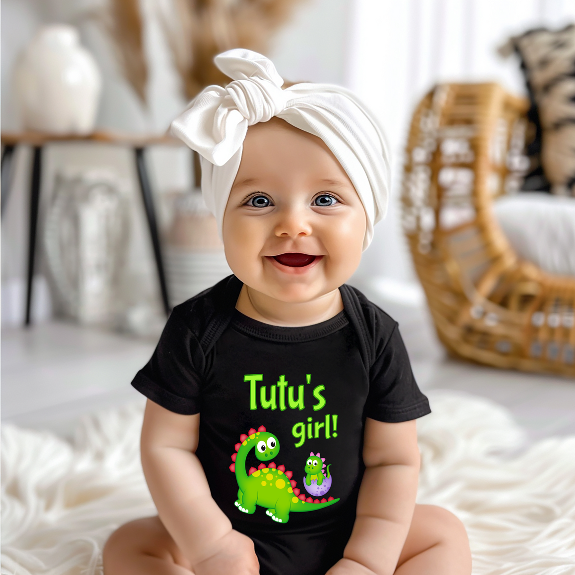 A smiling baby girl wearing a black t-shirt with a dinosaur graphic and the text "Tutu's girl!"