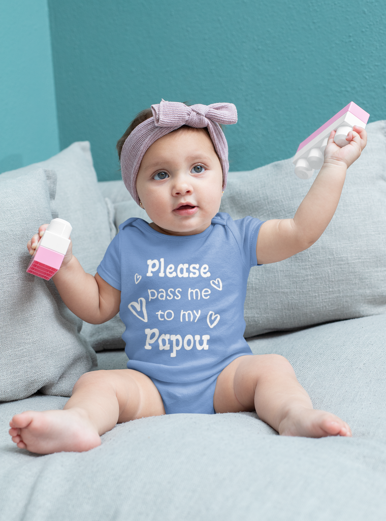 A baby girl wearing a blue onesie with the text "Please pass me to my Papou" is sitting on a gray couch, holding a pink toy.
