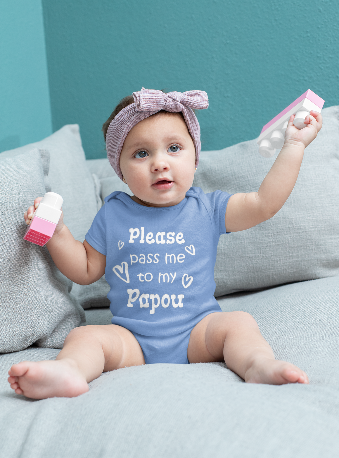 A baby girl wearing a blue onesie with the text "Please pass me to my Papou" is sitting on a gray couch, holding a pink toy.