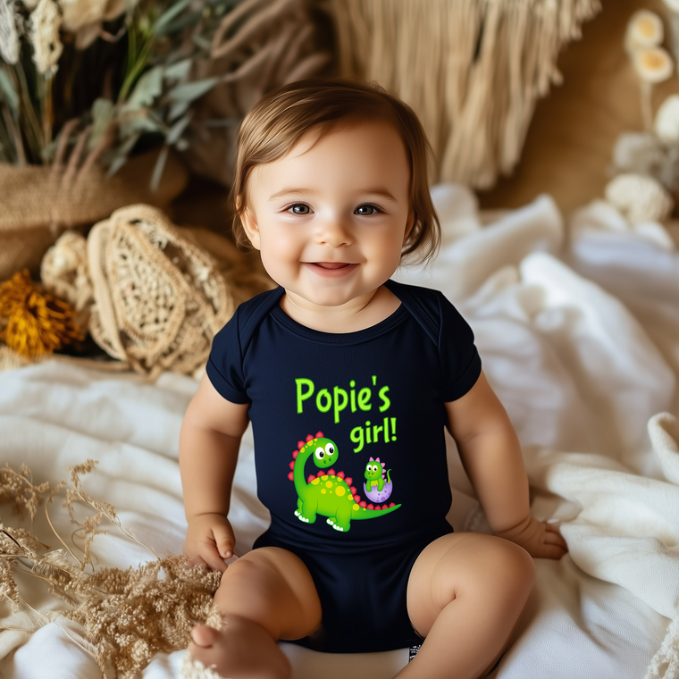 A smiling baby wearing a black shirt with a dinosaur graphic and the text "Poppy's girl!"