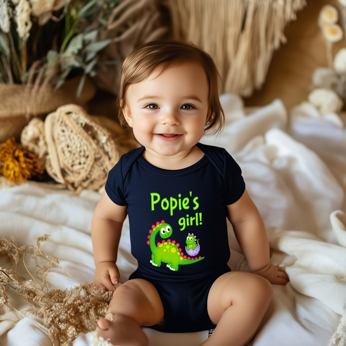 A smiling baby wearing a black shirt with a dinosaur graphic and the text "Poppy's girl!"