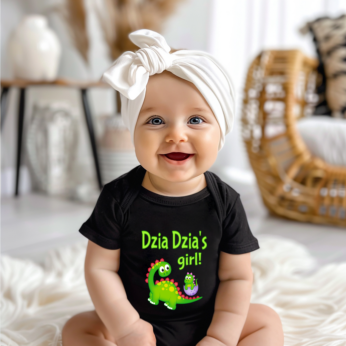 A smiling baby girl wearing a black t-shirt with a dinosaur graphic and the text "Dzia Dzia's girl!" is sitting on a white surface.