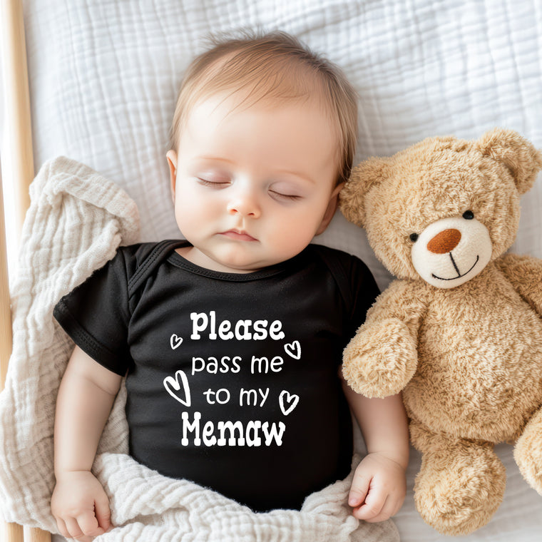 A sleeping baby wearing a black shirt with the text "Please pass me to my Memaw" is lying next to a teddy bear on a white blanket.