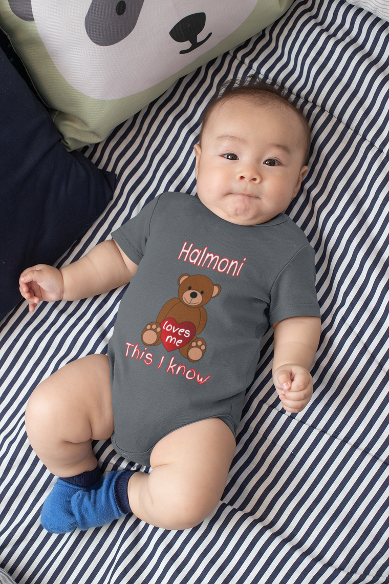 A baby wearing a grey onesie with a teddy bear graphic and the text "This I know" lying on a striped bed.