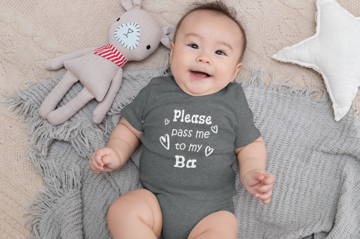 A smiling baby wearing a gray onesie with the text "Please pass me to my Ba" lies on a gray blanket next to a stuffed animal.
