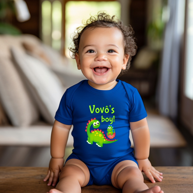 A smiling baby boy wearing a blue onesie with a dinosaur graphic and the text "VovŽ's boy!" is sitting on a wooden surface.