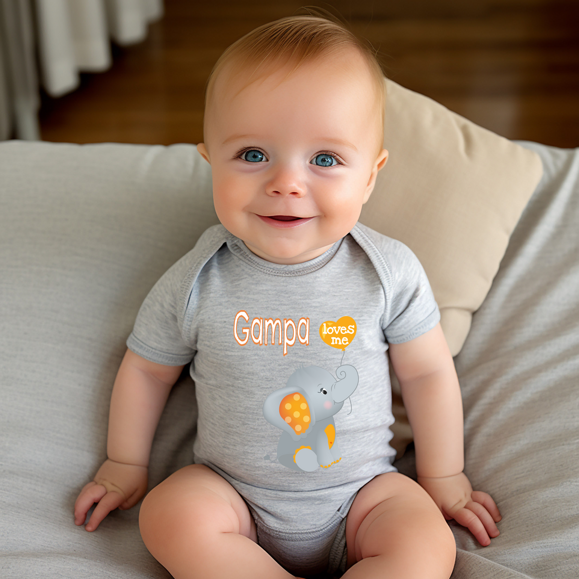 A smiling baby boy wearing a gray onesie with an elephant design sits on a couch.
