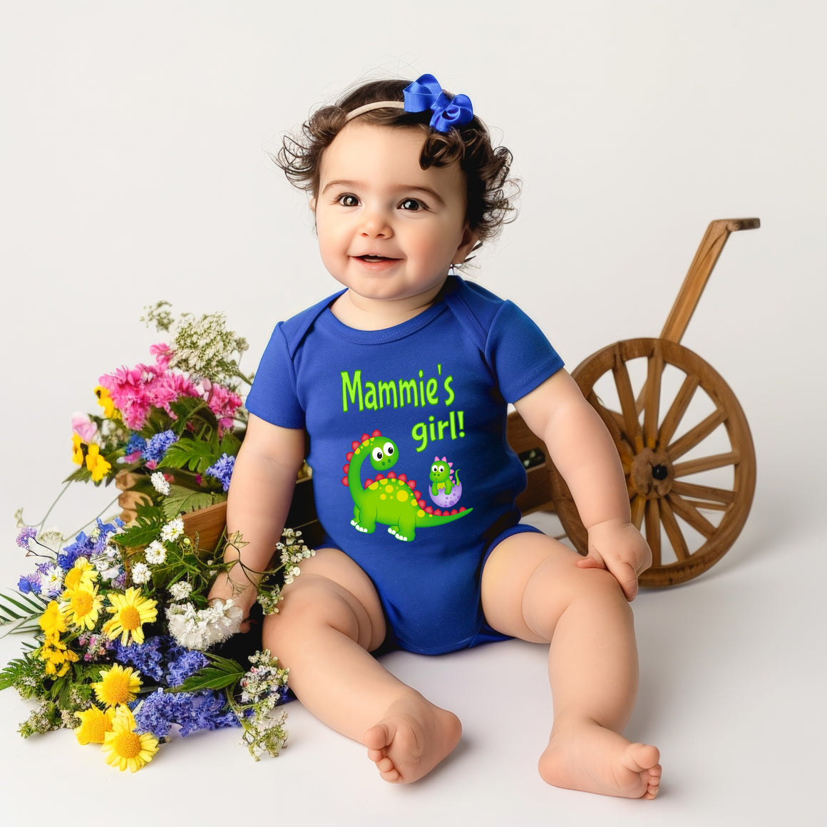 A smiling baby girl wearing a blue onesie with a dinosaur on it, sitting next to a wooden wagon filled with colorful flowers.